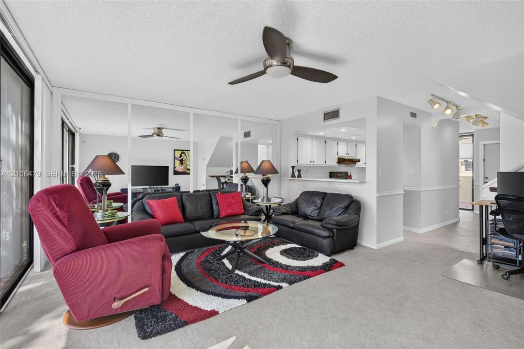Living area featuring a ceiling fan, wall-to-wall mirrored panels, carpeted flooring, and a pass-through opening to the kitchen