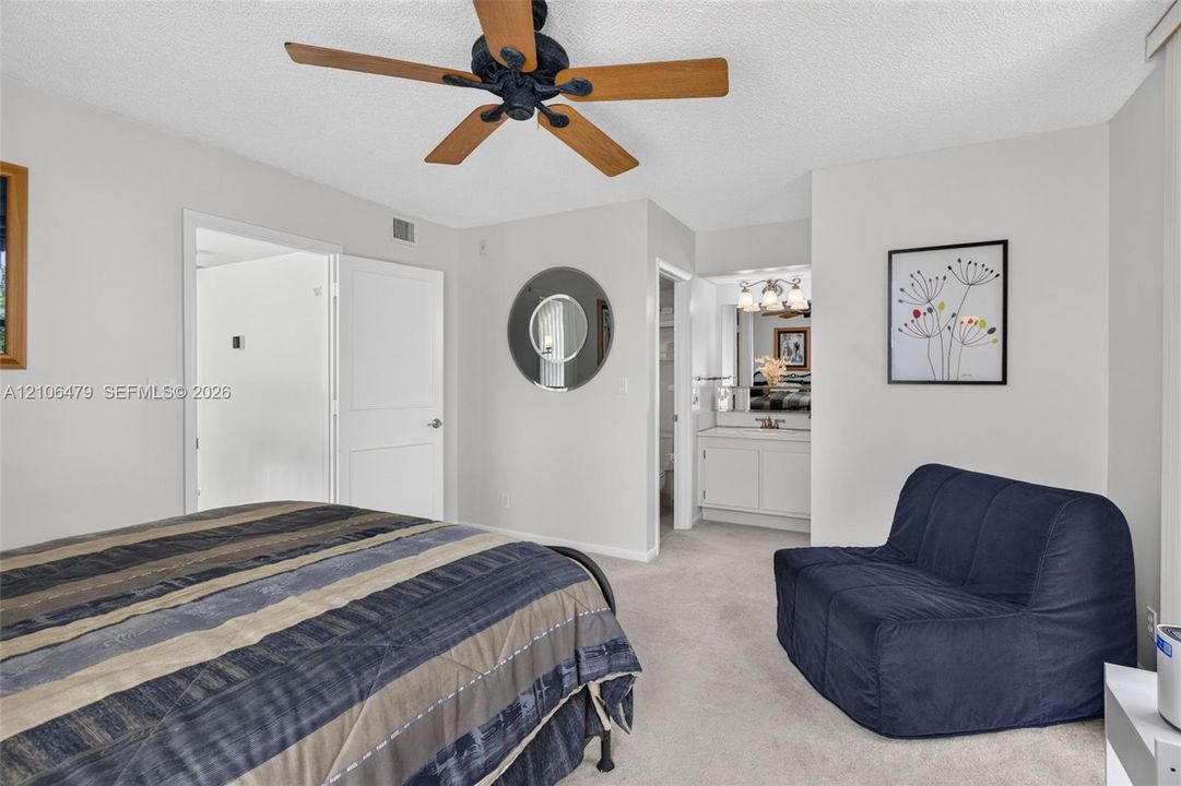Neutral interior space featuring carpeted flooring, a ceiling fan with wood-finish blades, a circular wall mirror, and a built-in vanity cabinet
