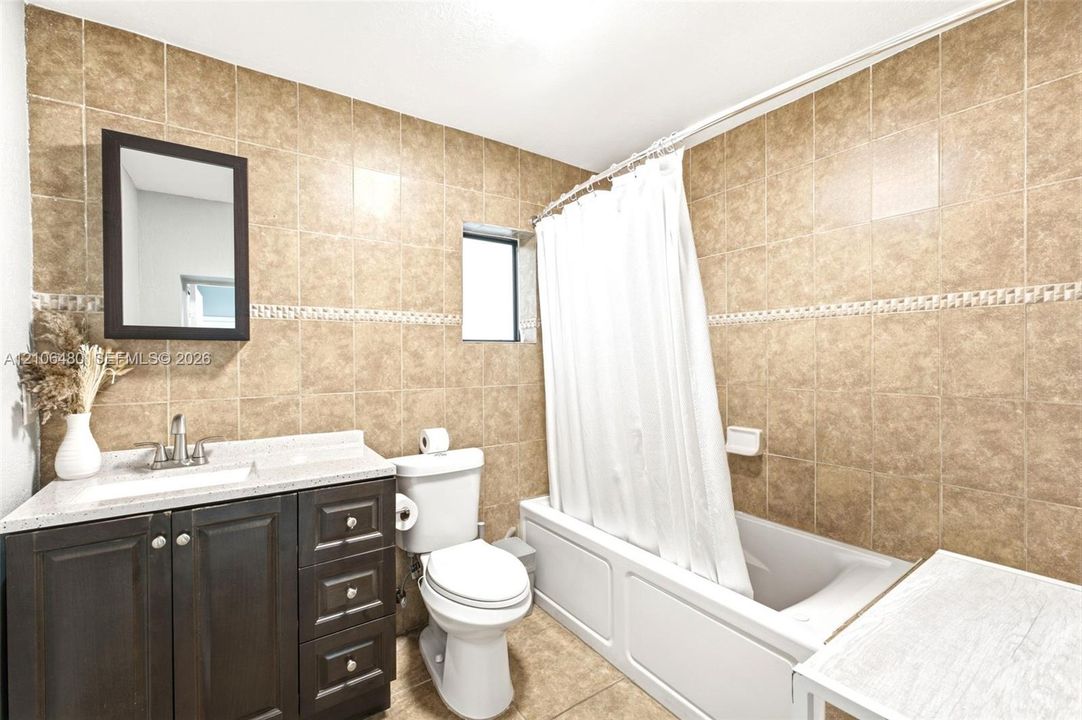 Bathroom featuring tile-surround walls with decorative border, dark wood-finish vanity with stone-composite countertop, undermount sink, and deep soaking tub with shower combination