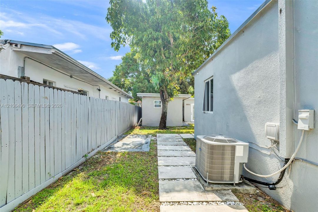 Exterior side yard featuring a white wood-panel fence, a concrete paver walkway, mature landscaping, and a detached outbuilding