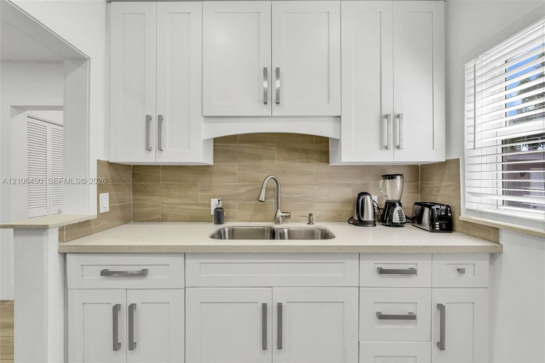 Shaker-style white cabinetry with brushed nickel hardware, neutral solid-surface countertops, horizontal tile backsplash, stainless steel dual-basin undermount sink, and pull-down faucet