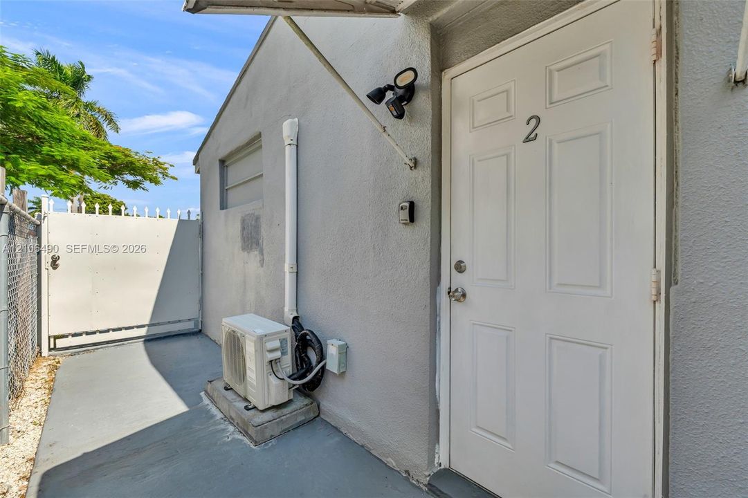 Stucco exterior featuring a six-panel entry door, exterior wall-mounted light fixture, and dedicated mini-split HVAC unit on a paved concrete pad