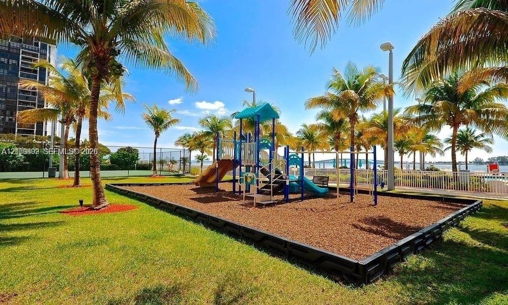 Outdoor recreational area featuring a playground structure with slides, a manicured lawn, established palm trees, and perimeter fencing