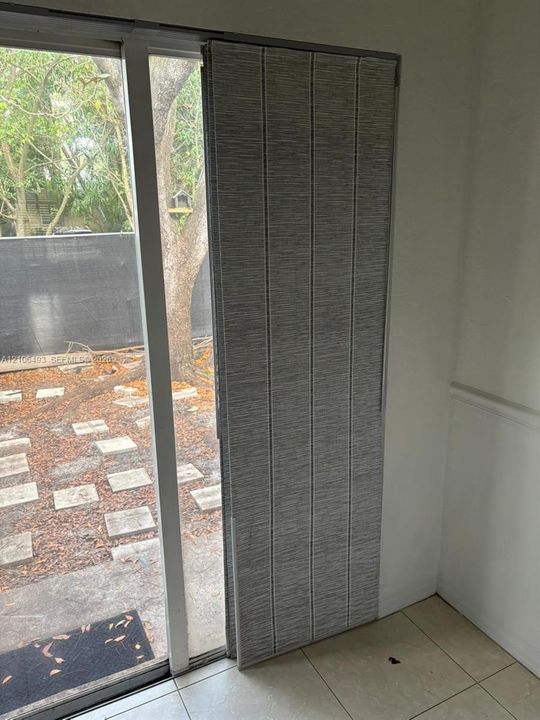 Large-format glass sliding door with white frame and floor-to-ceiling textured panel blinds, featuring ceramic tile flooring and outdoor access to a patio with stone pavers