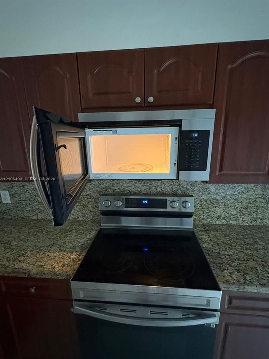 Stainless steel over-the-range microwave and electric range, dark wood-finish cabinetry, and stone-surface countertops with matching backsplash