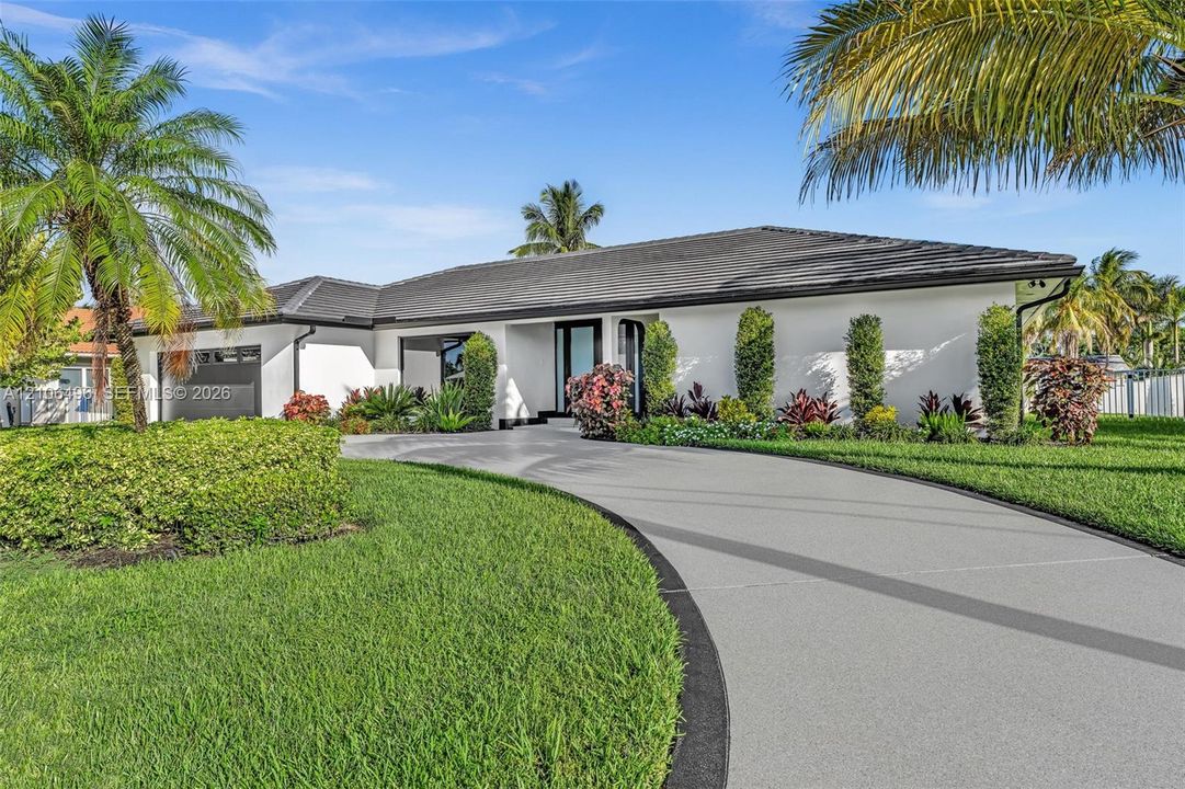 Single-story residence featuring a low-pitched tiled roof, smooth stucco exterior, circular concrete driveway with decorative border, and established tropical landscaping