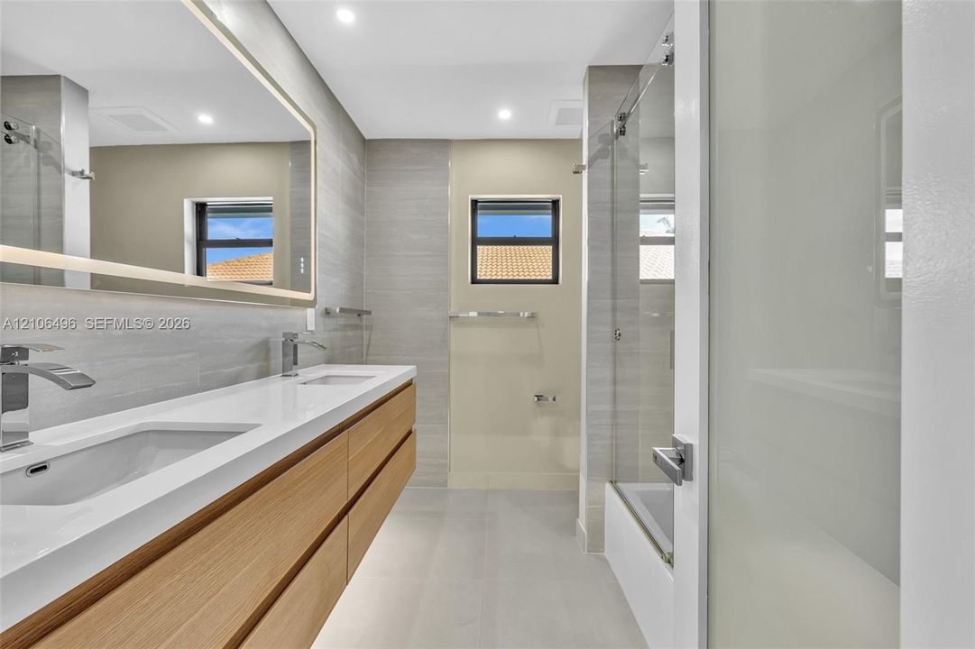 Floating double vanity with a stone countertop and dual undermount sinks, illuminated mirror, wood-finish cabinetry, and a glass-enclosed shower featuring textured wall tile