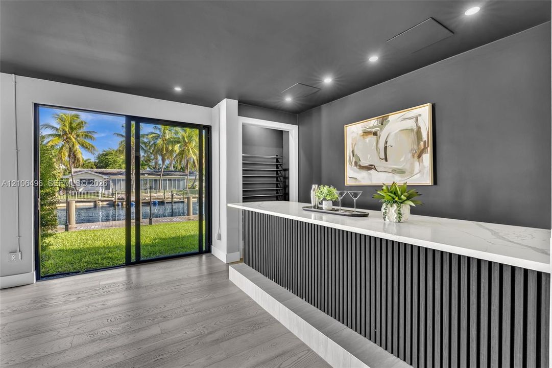 Built-in bar featuring a stone-surface countertop, slatted wood-finish base, recessed ceiling lighting, and sliding glass doors with water and landscape access
