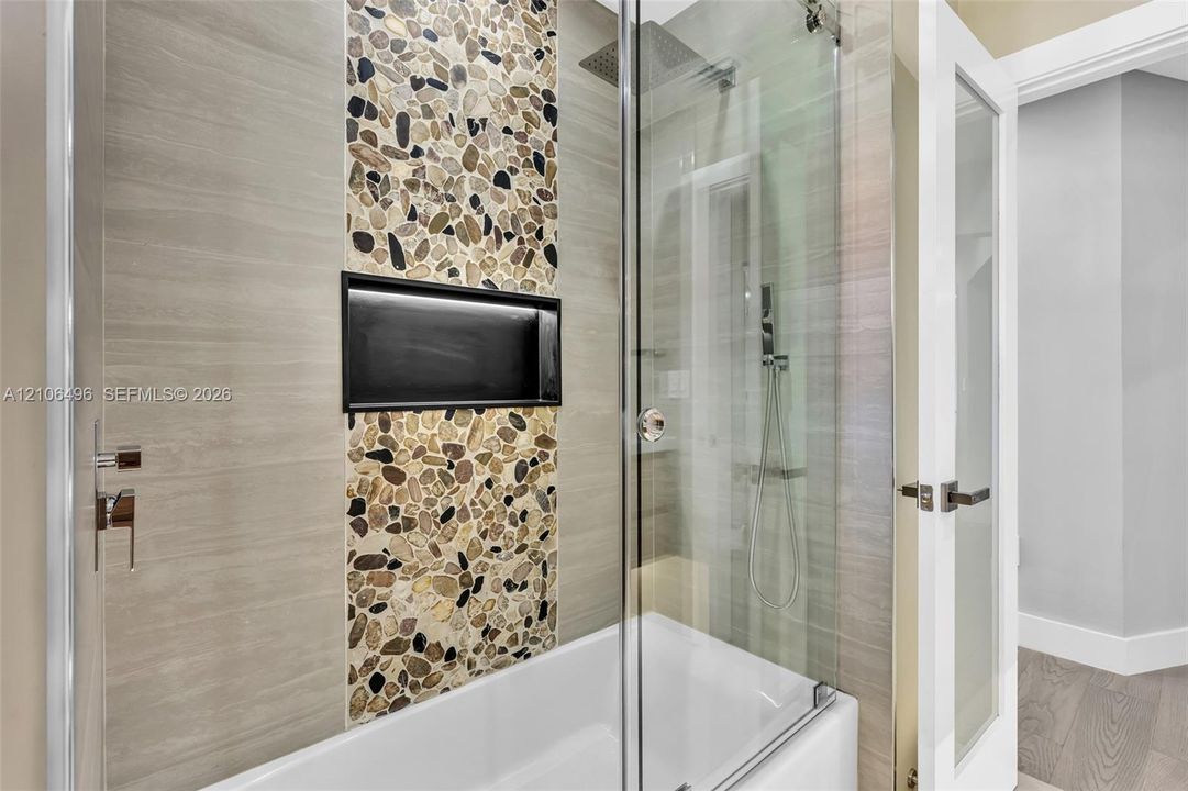Shower and tub combination featuring horizontal tile surrounds, a decorative vertical pebble-tile inlay, integrated black-framed shower niche, glass shower screen, and rainfall showerhead