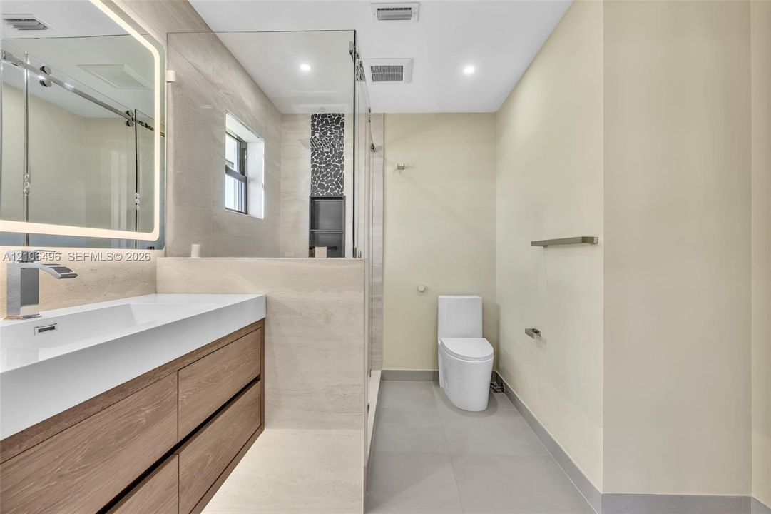 Floating wood-finish vanity with an integrated rectangular sink, backlit vanity mirror, glass-enclosed shower with decorative stone-look inlay, and large-format tile flooring