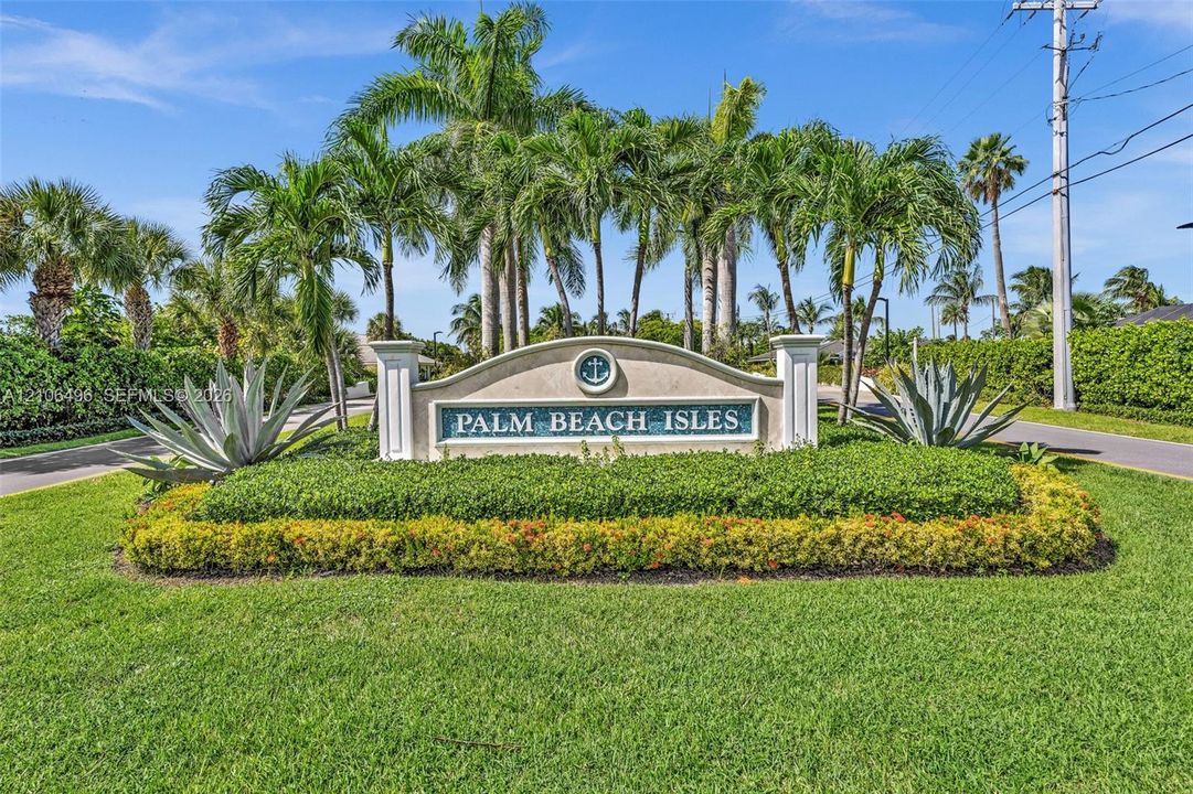 Palm Beach Isles community entrance featuring a stone monument sign, manicured lawn, established palm trees, and structural landscape hedging