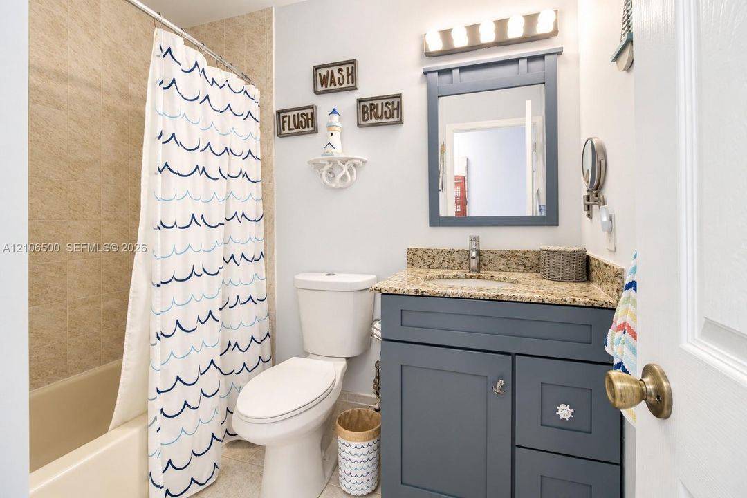 Blue vanity cabinet with a speckled stone countertop, integrated sink, matching framed mirror, and a tub-shower combination with textured surround tile