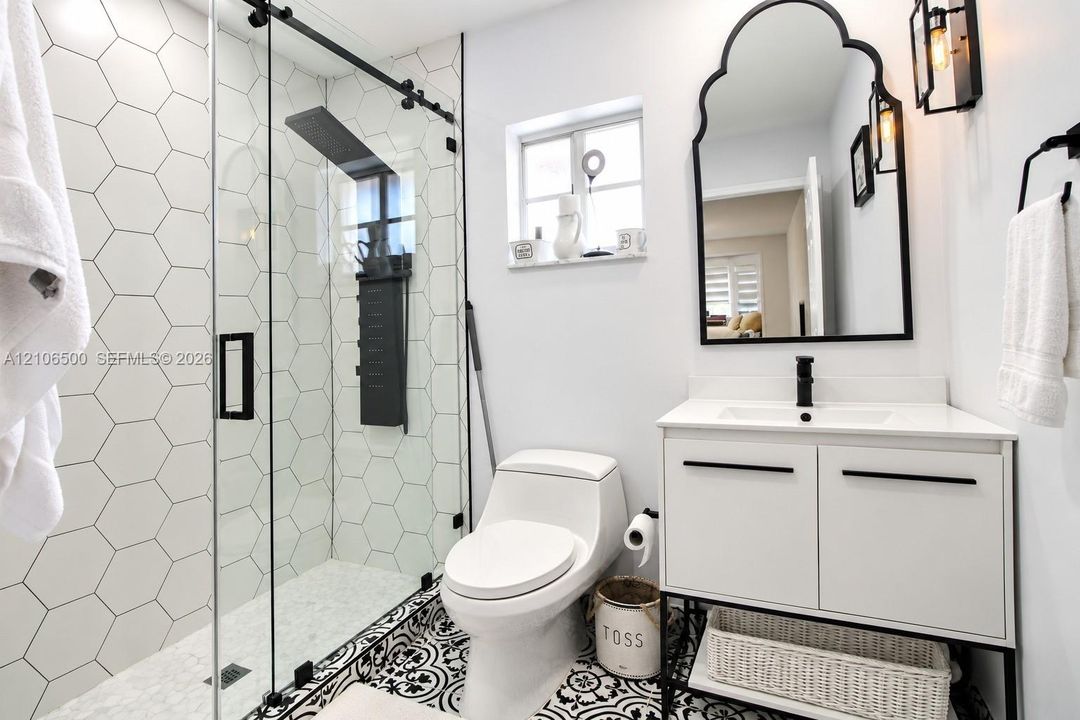 Walk-in shower with hexagon wall tile, sliding glass door, and rainfall showerhead; vanity featuring a rectangular sink, matte black hardware, and an arched mirror