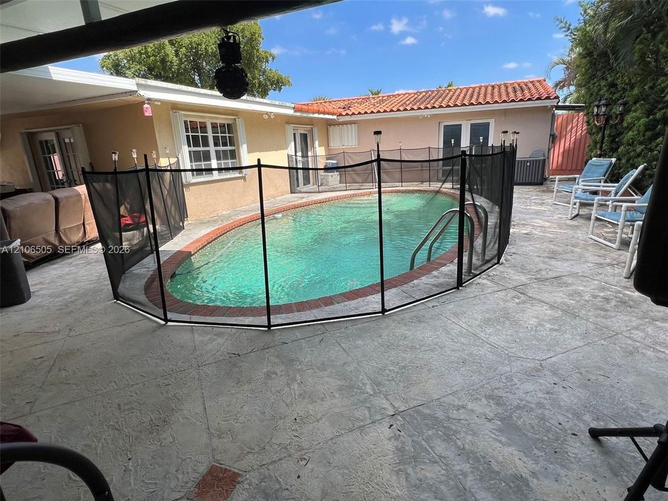Oval swimming pool with brick coping, enclosed by a removable mesh safety fence and surrounded by an expansive concrete patio