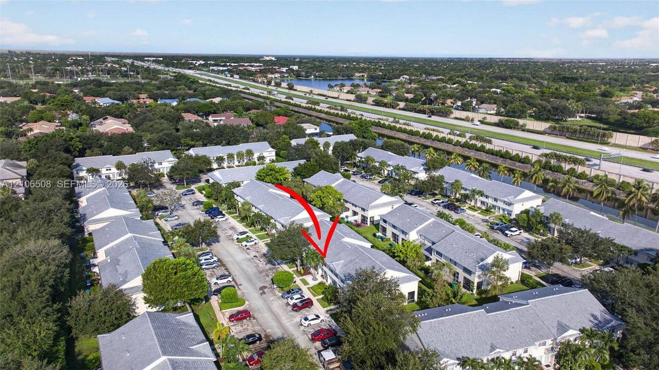 Aerial perspective of multi-family complex featuring light-colored shingled roofing, paved surface parking, and mature landscape elements