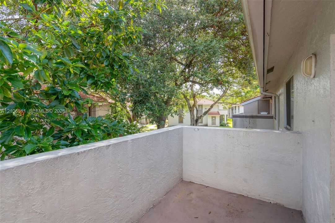 Covered balcony with textured stucco walls, solid wall railing, and an attached exterior light fixture, surrounded by mature trees