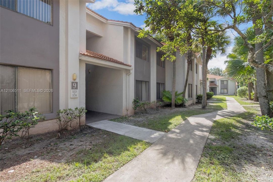 Multi-story exterior featuring a neutral stucco facade, terracotta-tiled roof accents, and concrete pathways surrounded by mature palms and landscaping
