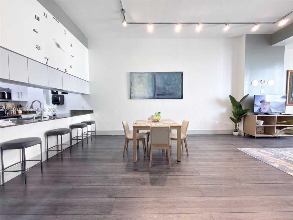 Open-concept interior featuring wood-finish flooring, track lighting, cabinetry with matte-finish upper doors, and a stone-surface breakfast bar