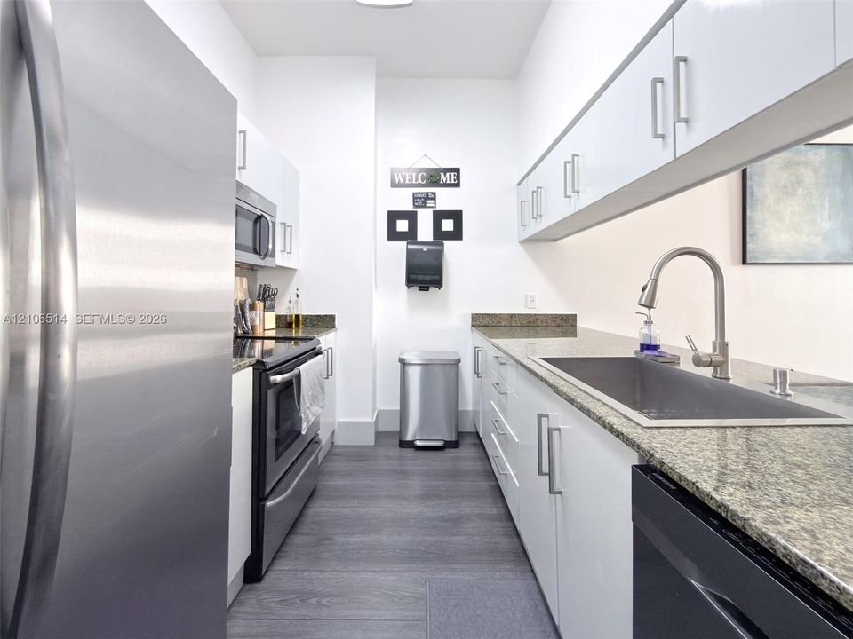 Galley-style kitchen featuring high-gloss white cabinetry, stone countertops, stainless steel appliances, and wood-finish flooring