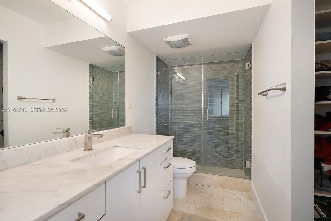 Bathroom featuring a white vanity with a stone countertop, an integrated rectangular sink, a full-width wall mirror, and a glass-enclosed shower with textured tile surround
