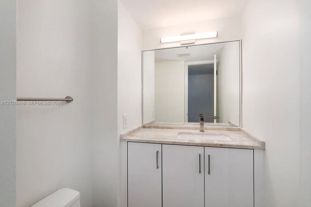 White vanity cabinetry with a stone-surface countertop, rectangular undermount sink, polished metal faucet, full-width mirror, and bar-style wall lighting