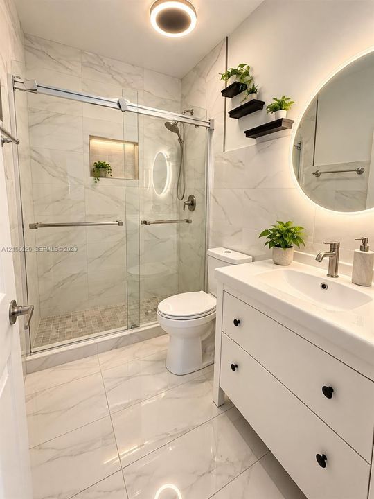 Bathroom featuring a sliding glass shower door, stone-finish tile surround, built-in recessed shower niche, vanity with integrated basin, and illuminated wall-mounted mirror