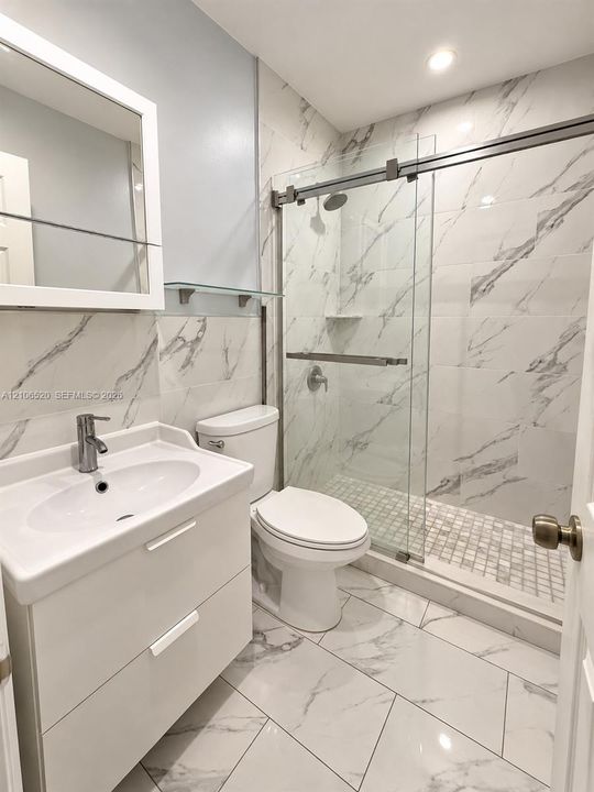 Bathroom featuring a white dual-drawer vanity with integrated sink, glass-enclosed shower with floor-to-ceiling tile, and porcelain-finish tiled flooring throughout