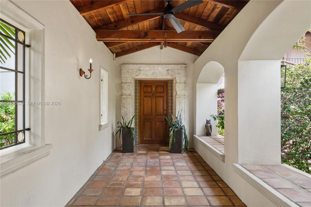 Covered entryway featuring terracotta tile flooring, exposed wood-beam vaulted ceiling, arched masonry openings, stone door surround with column detailing, and built-in masonry seating