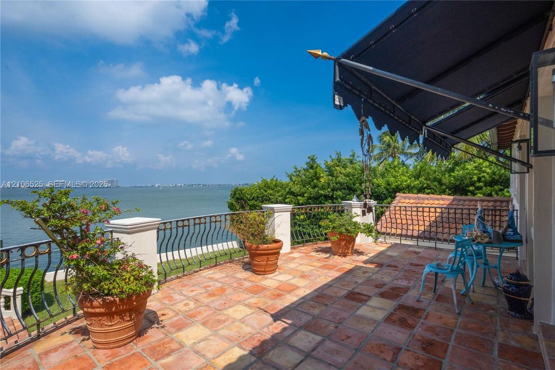 Open-air terrace featuring terra-cotta tile flooring, ornate metal railing, retractable fabric awning, and unobstructed waterfront scenery