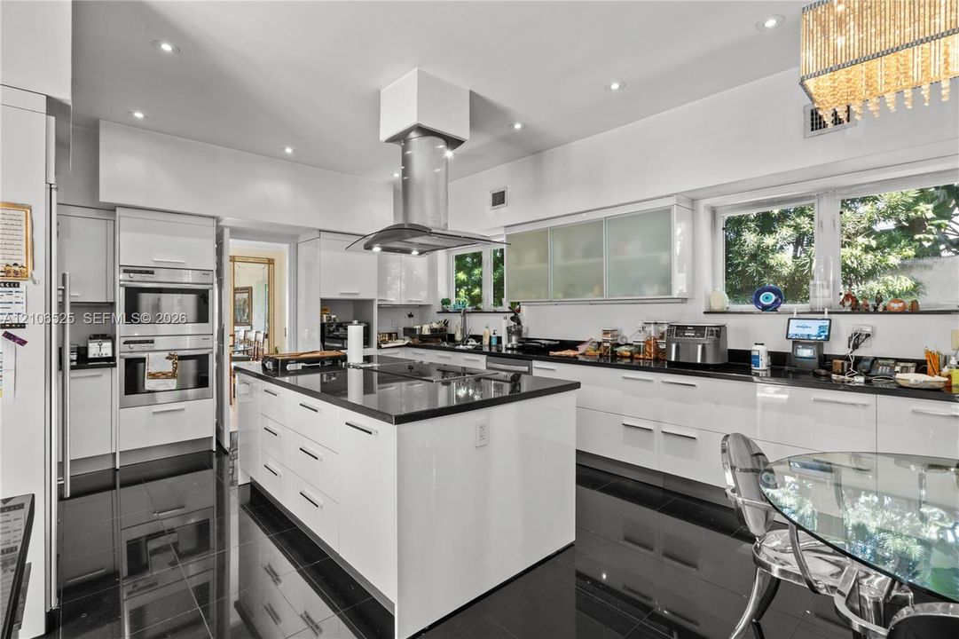 Modern kitchen featuring high-gloss cabinetry, a central island with a stone-finish countertop, stainless steel double wall ovens, a ceiling-mounted stainless steel range hood, and polished tile flooring