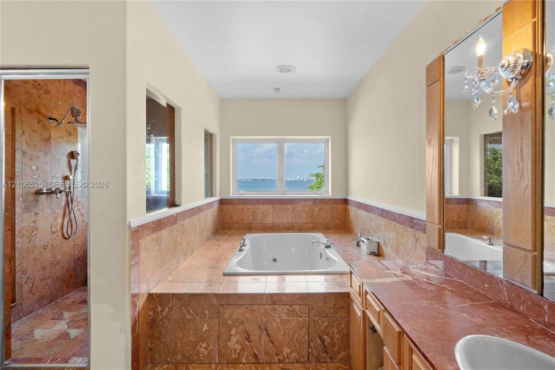 Bathroom featuring a built-in soaking tub, natural stone tile surround, walk-in shower with stone tile walls, stone-topped vanity, and wall-mounted light fixtures