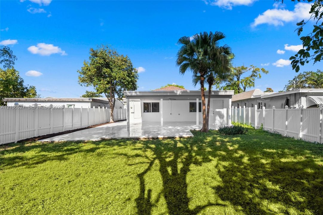 Rear exterior featuring a covered patio, paved hardscaping, white vinyl privacy fencing, and mature palm trees