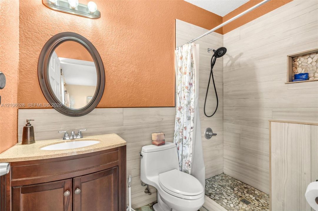 Bathroom featuring a wooden vanity with a stone countertop, an oval wall-mounted mirror, wood-finish wall tile, a stone-tiled shower floor, and a built-in shower niche