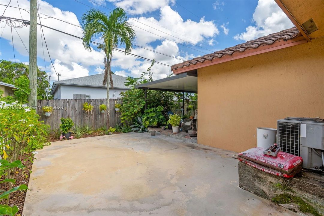 Exterior space featuring a covered patio, concrete hardscaping, wood-style fencing, and established tropical landscaping