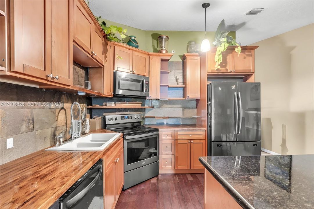 Kitchen featuring wood cabinetry, butcher block countertops, stone-tile backsplash, stainless steel appliances, and a dark stone-surface island