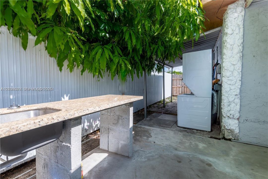 Outdoor utility area featuring a covered concrete patio, stone-finish countertop with an integrated metal sink, stacked laundry unit, and metal privacy fencing