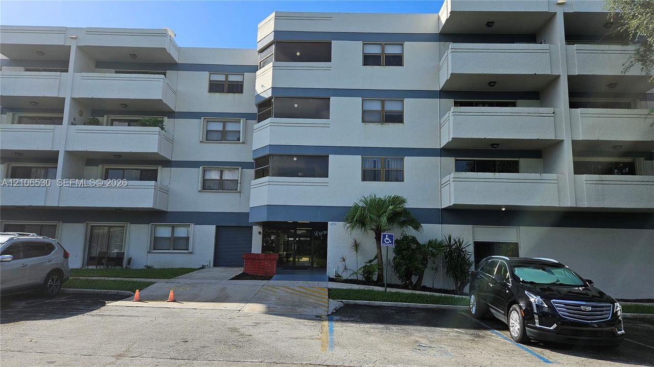 Multi-story residential building exterior featuring a concrete facade with horizontal detailing, individual private balconies, glass-pane windows, and paved surface parking