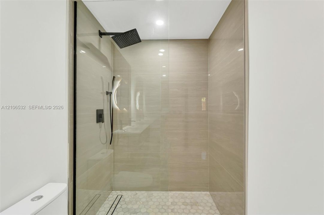 Walk-in shower featuring floor-to-ceiling large-format tile walls, glass enclosure, black rainfall showerhead with handheld attachment, and hexagonal tile flooring