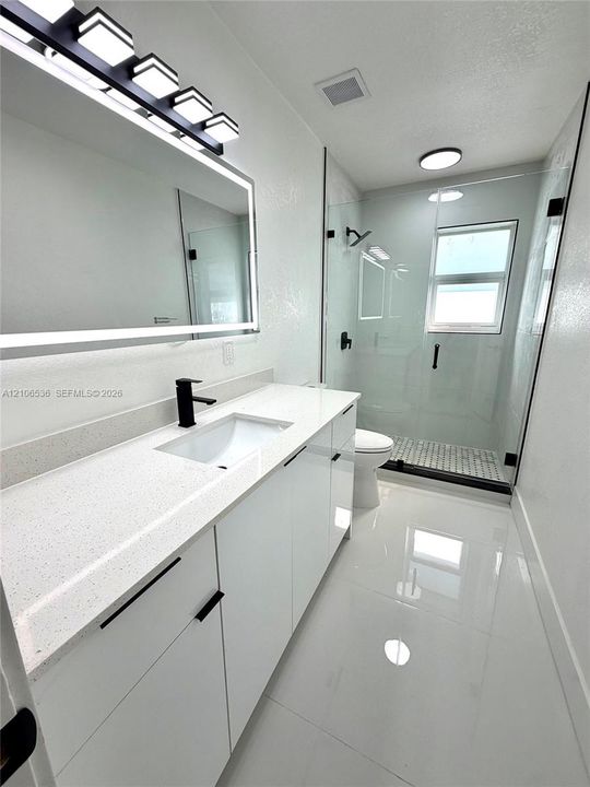 Modern vanity featuring white cabinetry, quartz countertop with an integrated basin, and a matte black faucet, illuminated by an LED-backlit mirror and five-light fixture, paired with a glass-enclosed shower featuring matte black hardware and light-toned flooring