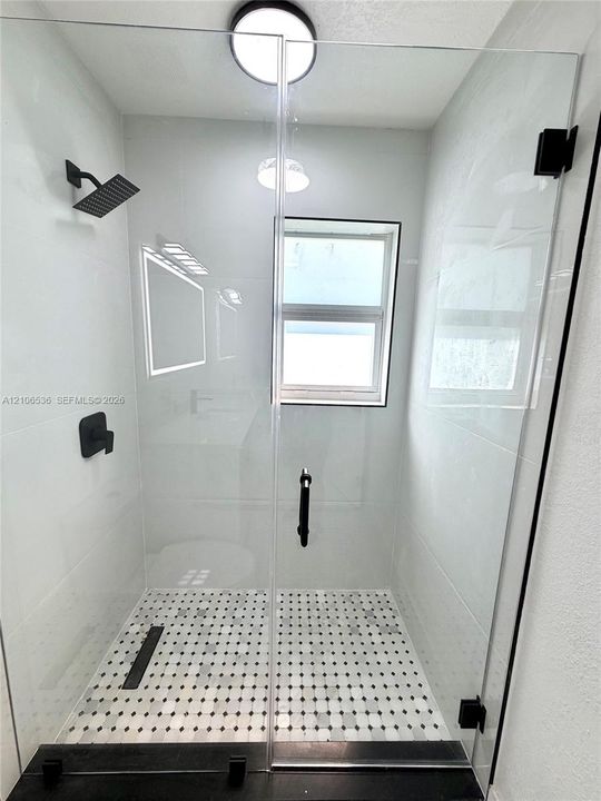 Walk-in shower featuring a frameless glass door, matte black fixtures, square rain-style showerhead, large-format wall tiling, and decorative mosaic floor tile with a linear drain
