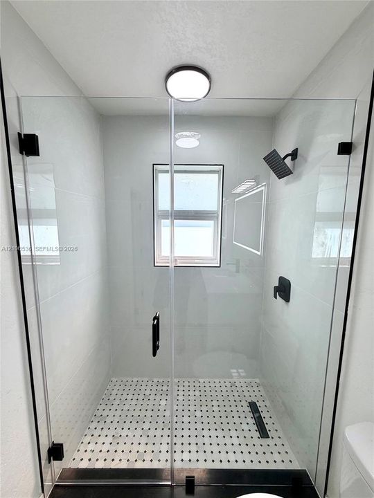 Walk-in shower featuring a frameless glass enclosure, matte black fixtures, white tiled walls, patterned floor tiling, and a recessed ceiling light