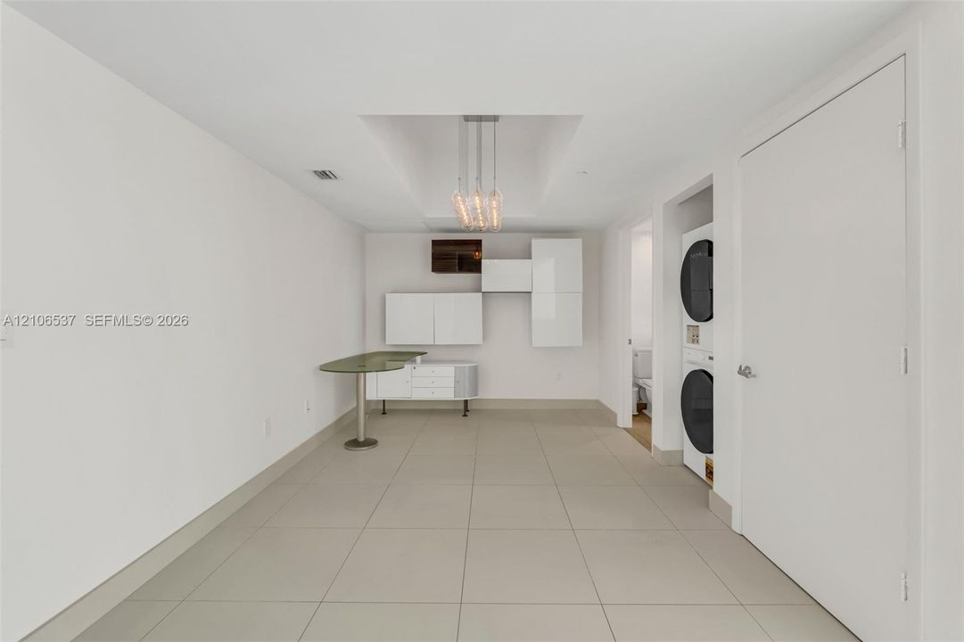 Modern utility space featuring large-format tile flooring, built-in glossy white cabinetry, an integrated stone-surface workstation, a tiered pendant light fixture, and a stacked washer and dryer unit