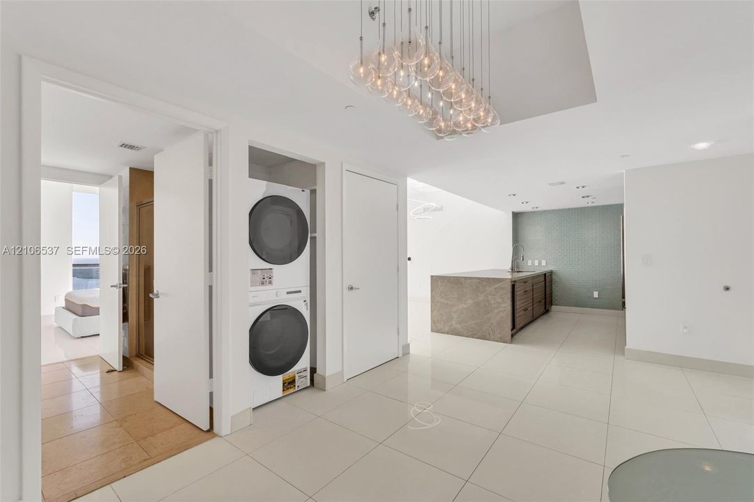 Large-format polished tile flooring, glass globe chandelier, stacked laundry unit, and kitchen island with stone-finish waterfall countertop