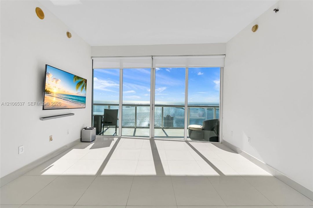 Floor-to-ceiling glass sliding doors, large-format tile flooring, and an attached balcony with metal railing, set against a waterfront backdrop