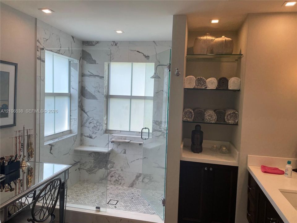 Walk-in shower featuring large-format patterned wall tile, a glass enclosure, a built-in stone bench, and recessed lighting, accompanied by built-in cabinetry with integrated shelving