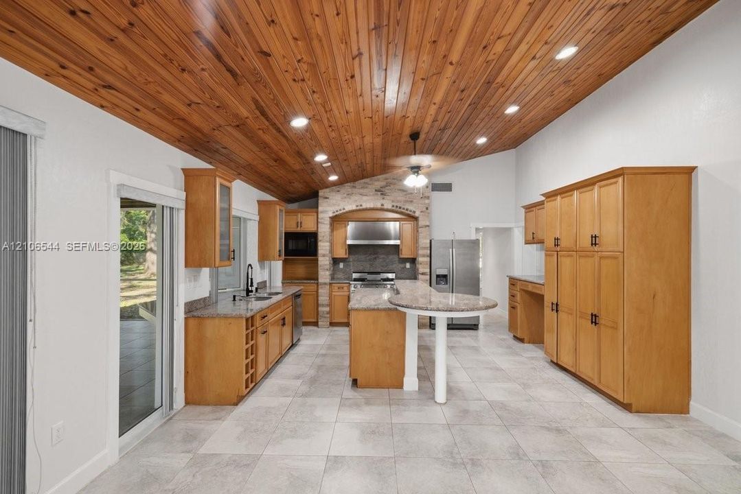 Vaulted wood-plank ceiling, natural stone-accented range alcove, custom cabinetry with built-in wine storage, stone-surface countertops, and large-format tile flooring