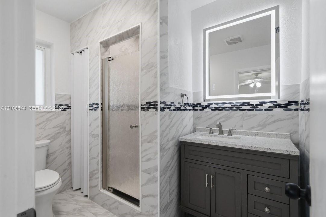 Bathroom featuring a dark wood-finish vanity with a stone-composite countertop, integrated rectangular sink, wall-mounted LED-backlit mirror, and shower enclosure with decorative tile inlay