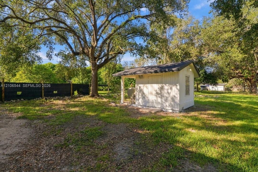 Detached storage structure featuring white siding, a shingled roof, a covered entry, and mature trees situated on a cleared lot with black perimeter fencing
