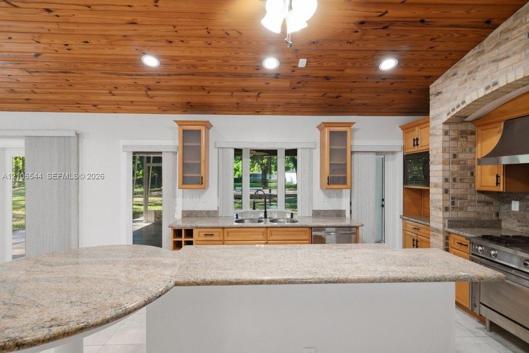 Kitchen featuring wood-plank cathedral ceilings, stone-surfaced kitchen island, custom cabinetry with glass-front uppers, brick-surround stove alcove, and stainless steel appliances
