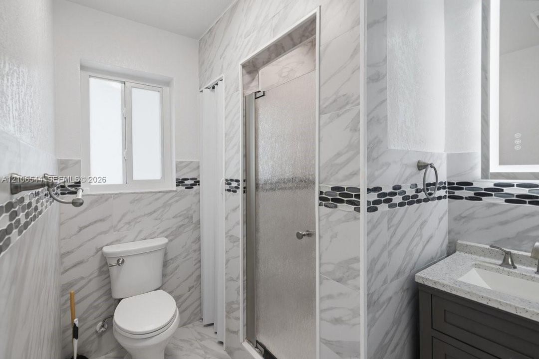 Bathroom featuring stone-finish wall tile with horizontal decorative accent inlay, enclosed shower with textured glass door, dark cabinetry with light-toned countertop, and integrated-light vanity mirror